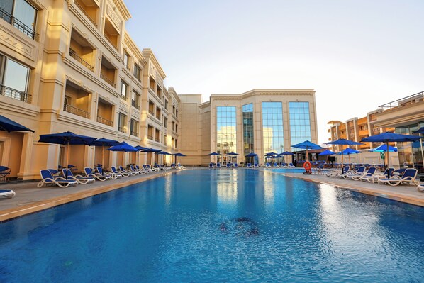 A swimming pool with lounge chairs and umbrellas, surrounded by multi-story buildings with balconies. - Helnan Mamoura Hotel & Events Center (Alexandria)