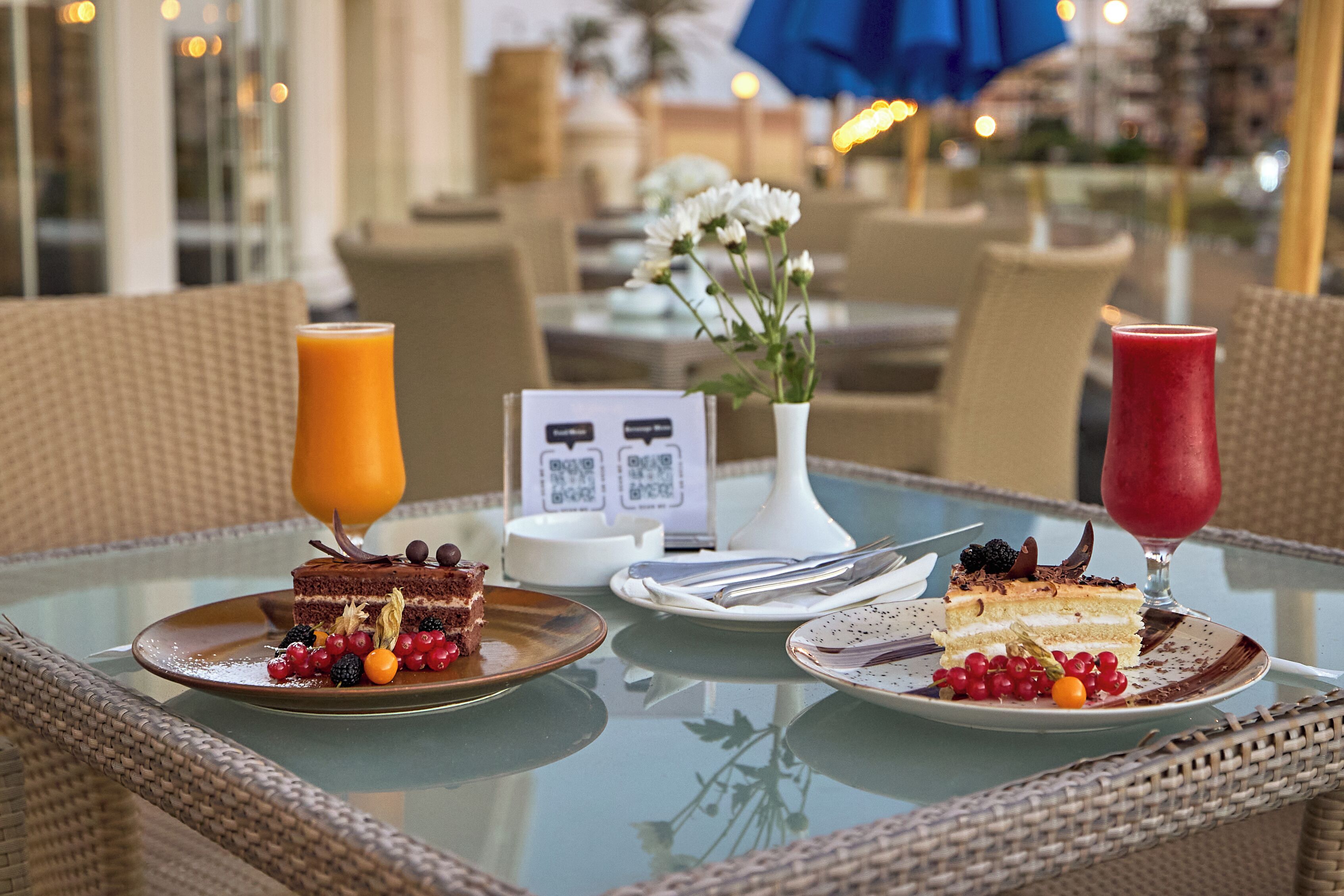 A glass table with two plates of cake, a glass of orange juice, and a glass of red drink.