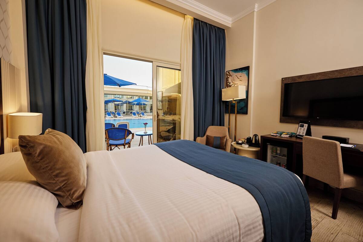 A hotel room with a large bed, a desk, a chair, a TV, and a view of a pool area.
