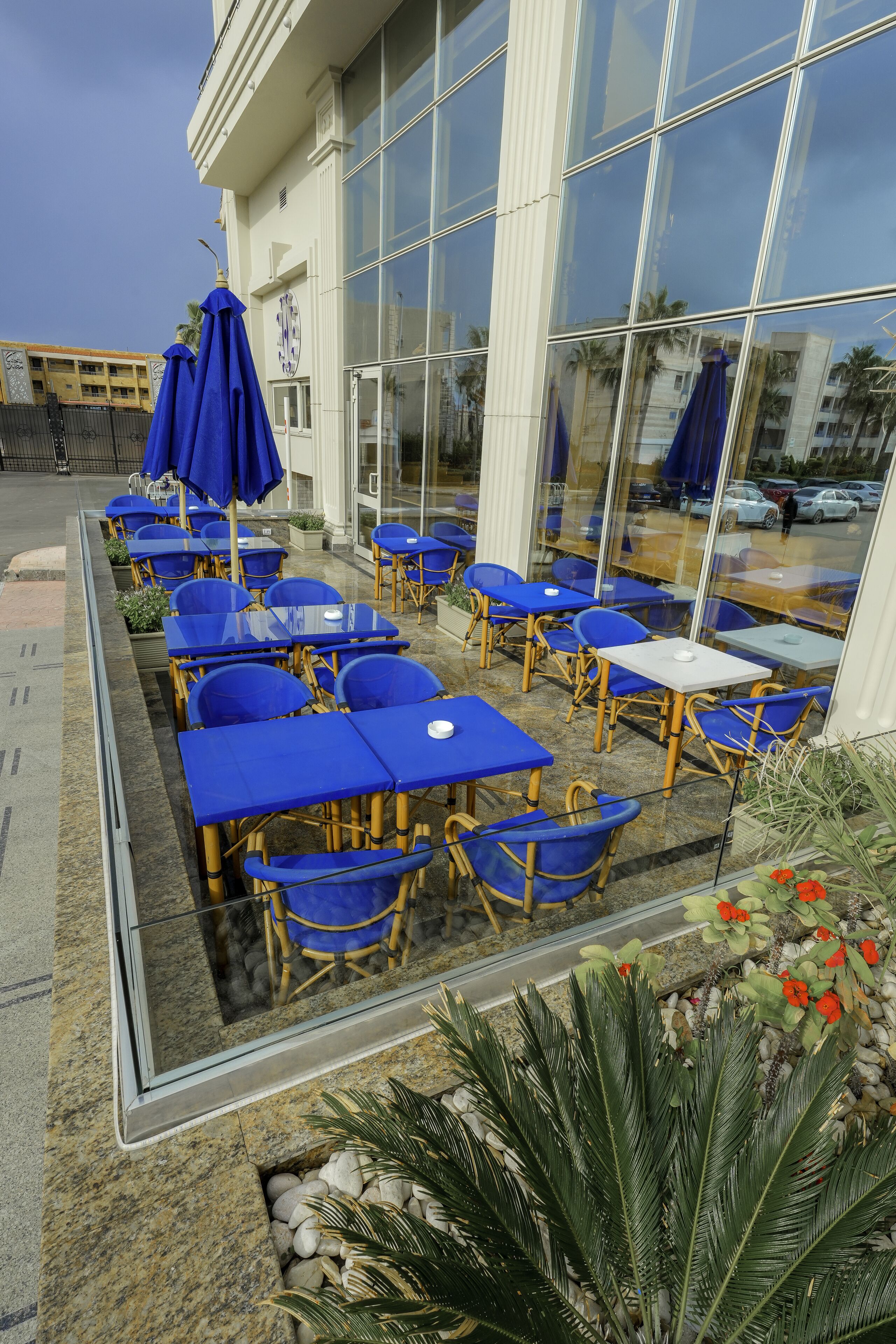 A seating area with tables and chairs, a clock, and a reflection of palm trees and cars in the glass facade.