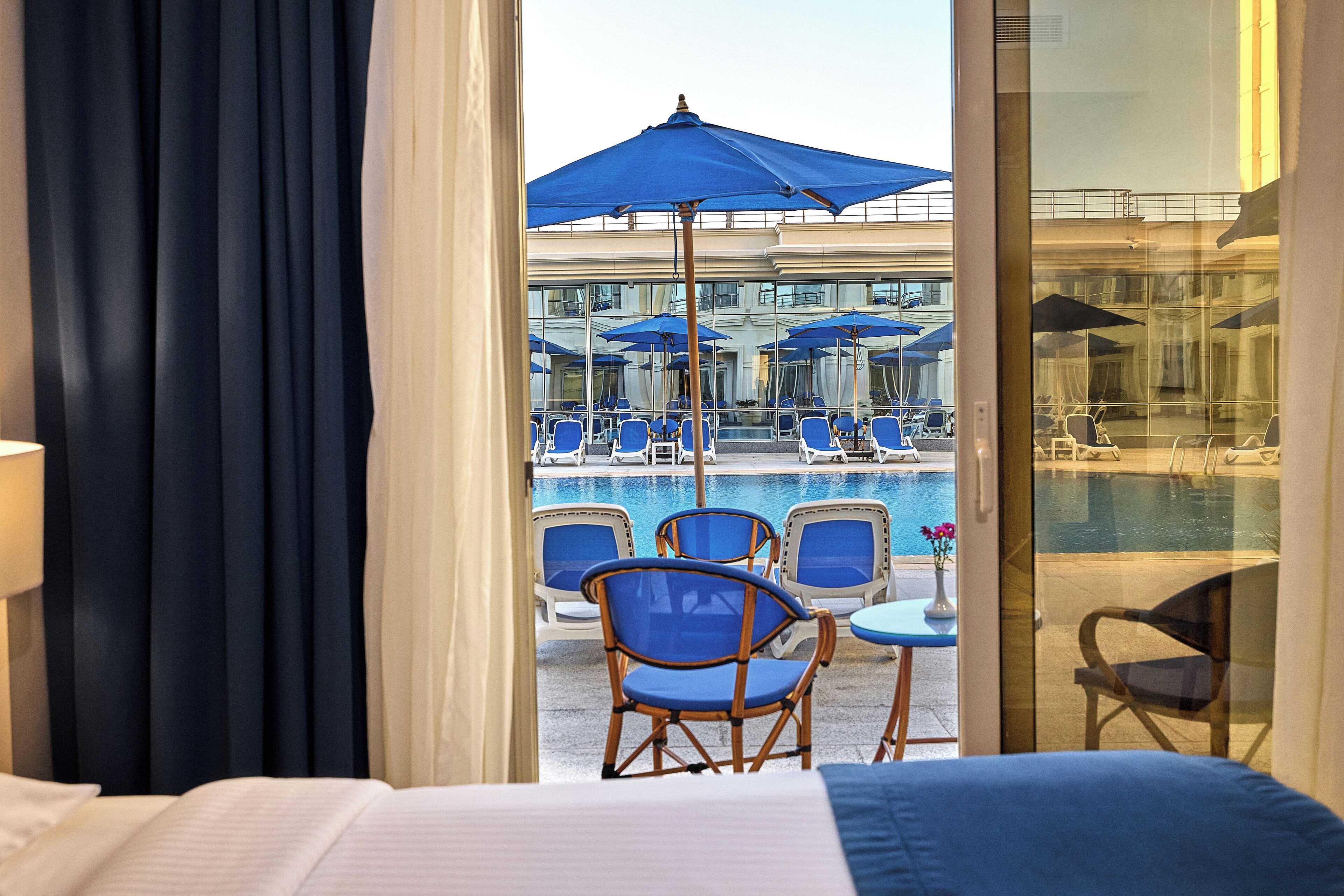 Une chambre d’hôtel avec un lit, donnant sur un espace piscine avec des chaises longues et des parasols, et une porte vitrée qui mène à la piscine.