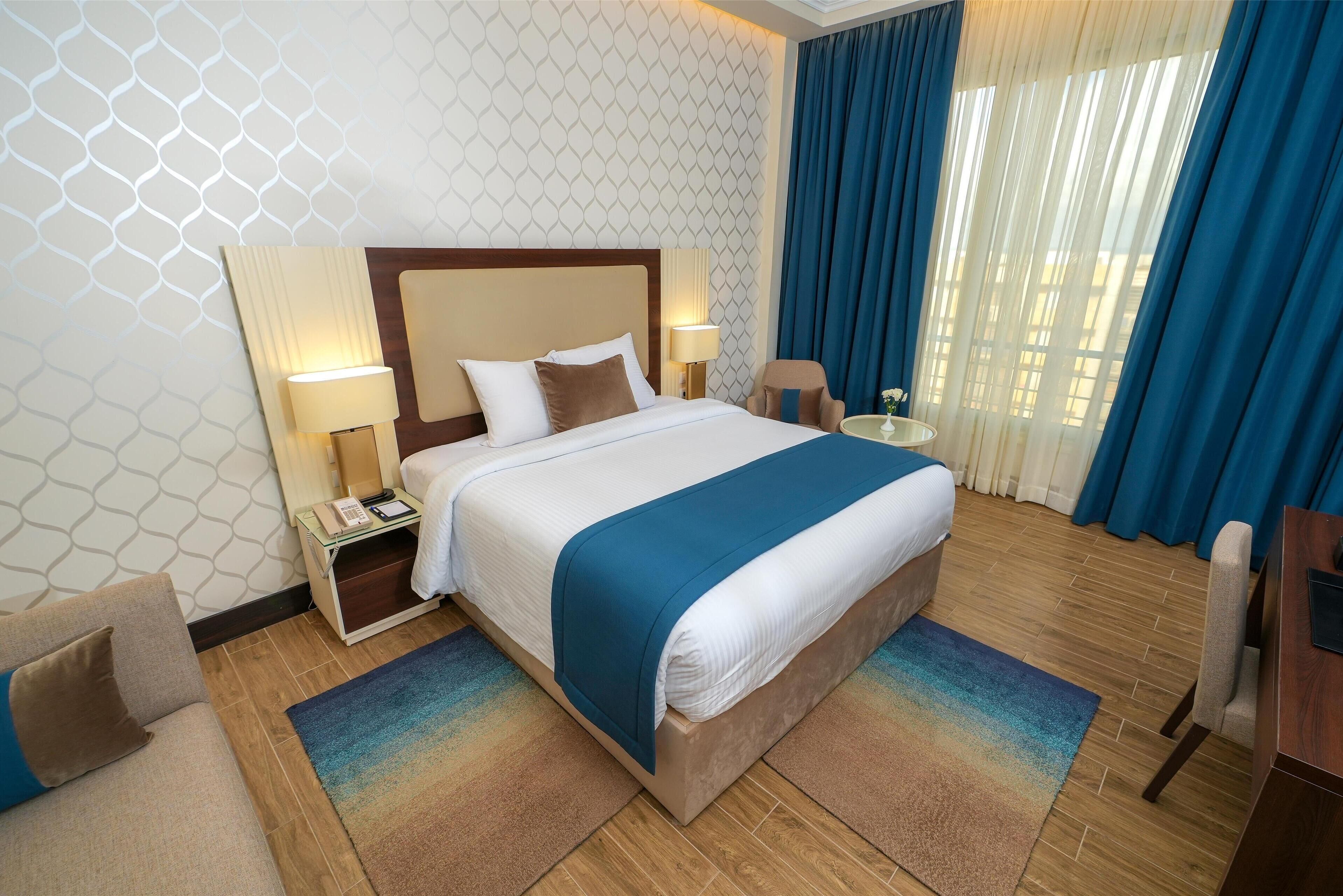 A hotel room with a large bed, two bedside tables with lamps, a chair, a small round table, and a view of the outdoors through blue curtains.