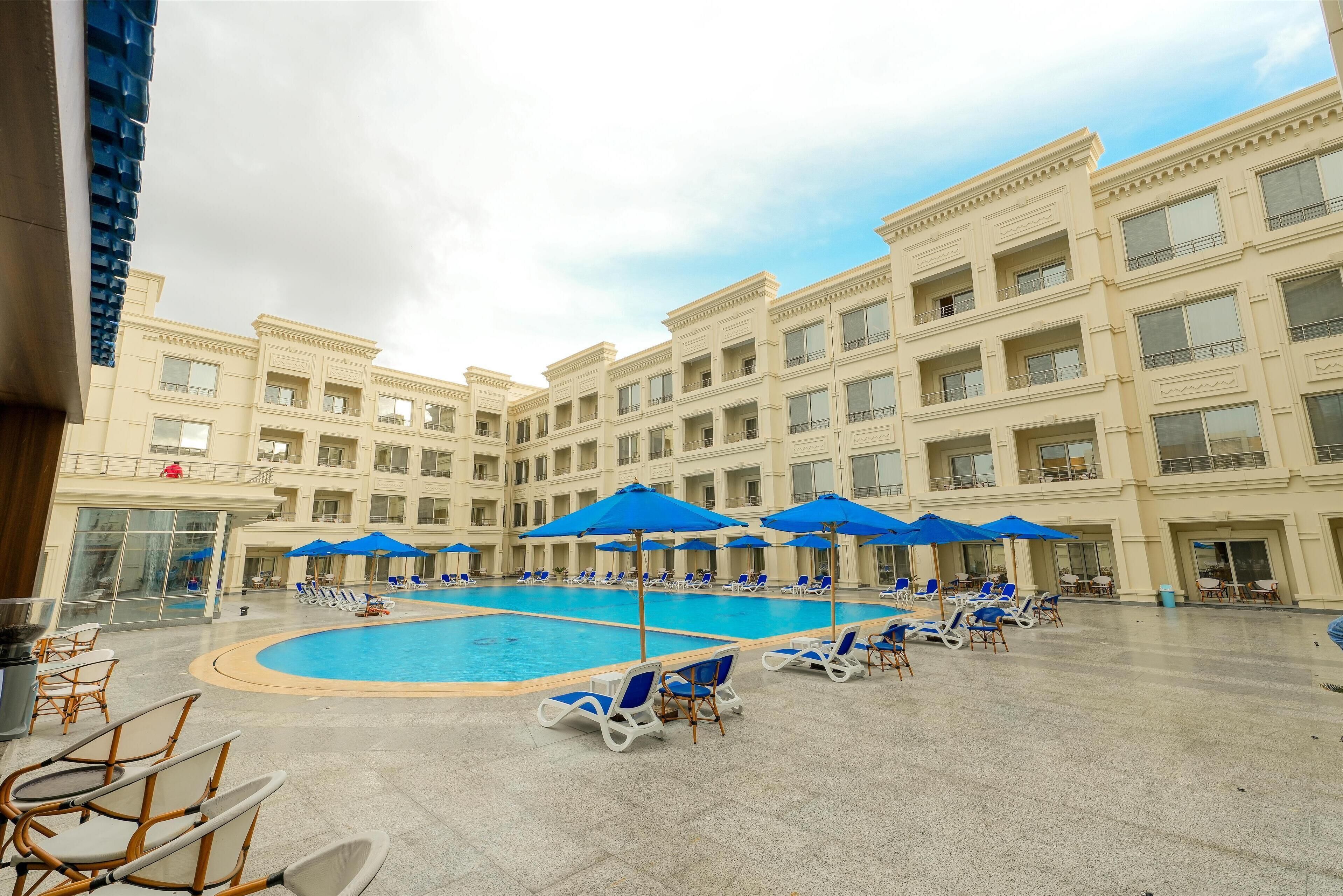 Un espace piscine d’hôtel avec des chaises longues et des parasols, entouré d’un immeuble à plusieurs étages doté de balcons.