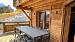 Een houten terras met een tafel en stoelen, grote ramen en uitzicht op een berg.