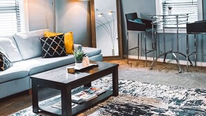 Un salon avec un canapé, une table basse et un tapis à motifs. On y trouve des luminaires au plafond, une horloge et une fenêtre avec des stores.