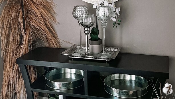 Une pièce avec un miroir rond, une console noire surmontée de deux bols en métal, une plante décorative et un mur orné d’un paysage marin peint.