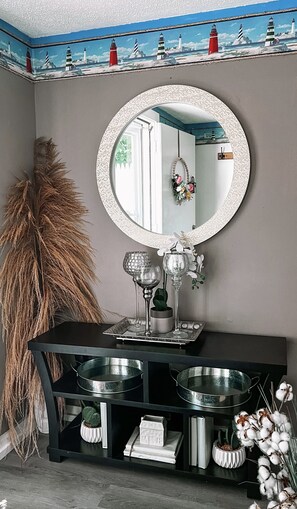 A room with a round mirror, a black console table with two metal bowls, a decorative plant, and a wall with a painted seascape.