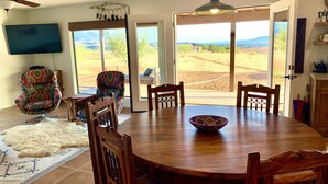 A dining area with a wooden table, four chairs, a patterned armchair, a television, and a view of the outdoors. - BirdSong Desert Retreat: Reconnect. Rejuvenate. (Nogales)