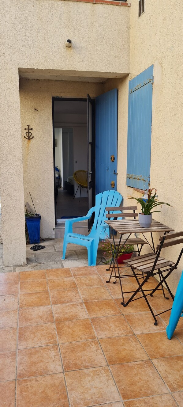 Een terras buiten met een tafel, stoelen, een plant in pot en een blauwe deur.