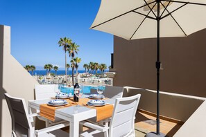 Une table à manger entourée de chaises blanches et surmontée d’un parasol beige, avec vue sur une piscine et des palmiers.