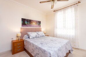 Une chambre à coucher avec un lit, des tables de chevet, un ventilateur de plafond, un tableau et une fenêtre avec des rideaux.