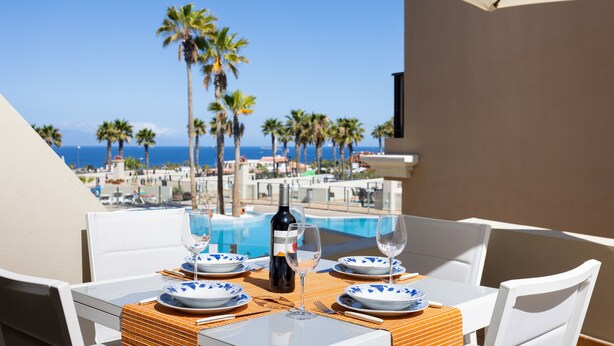 A table set for two with a bottle of wine, glasses, and plates, overlooking a pool and palm trees. - Apartment El Faro for 3 people in the south of Tenerife (Palm-Mar)