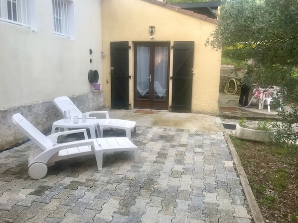 Un patio con sillones de plástico blanco y una mesa, una puerta con cortina, y un pequeño jardín con una manguera y una silla.