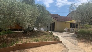 Una propiedad residencial con un camino pavimentado que conduce a una casa con techo de tejas rojas. Hay sillas al aire libre y un pequeño jardín con plantas y una jardinera de madera.