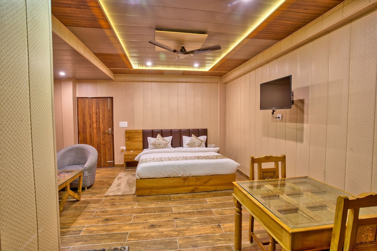 A hotel room with a wooden ceiling, a bed with white bedding, a television mounted on the wall, and a glass table.