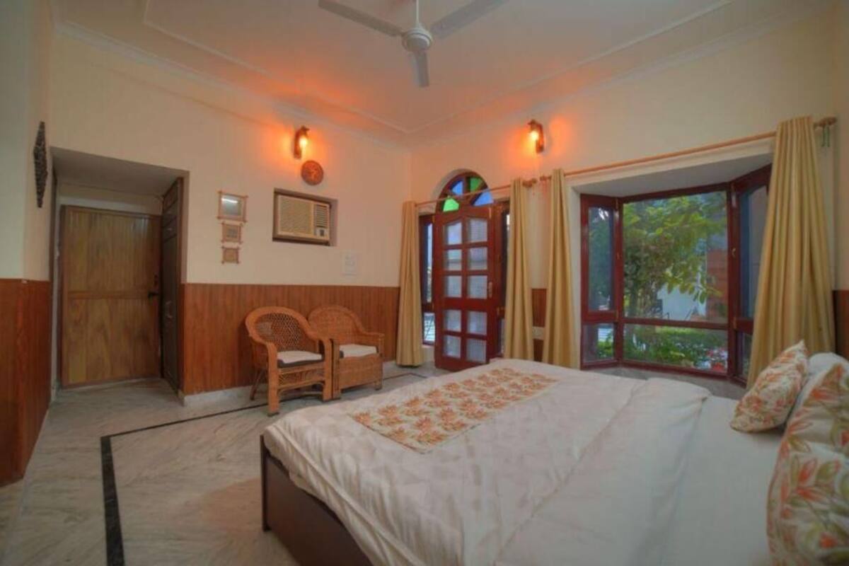 A bedroom with a bed, two chairs, a window with curtains, a door, and wall-mounted lights.
