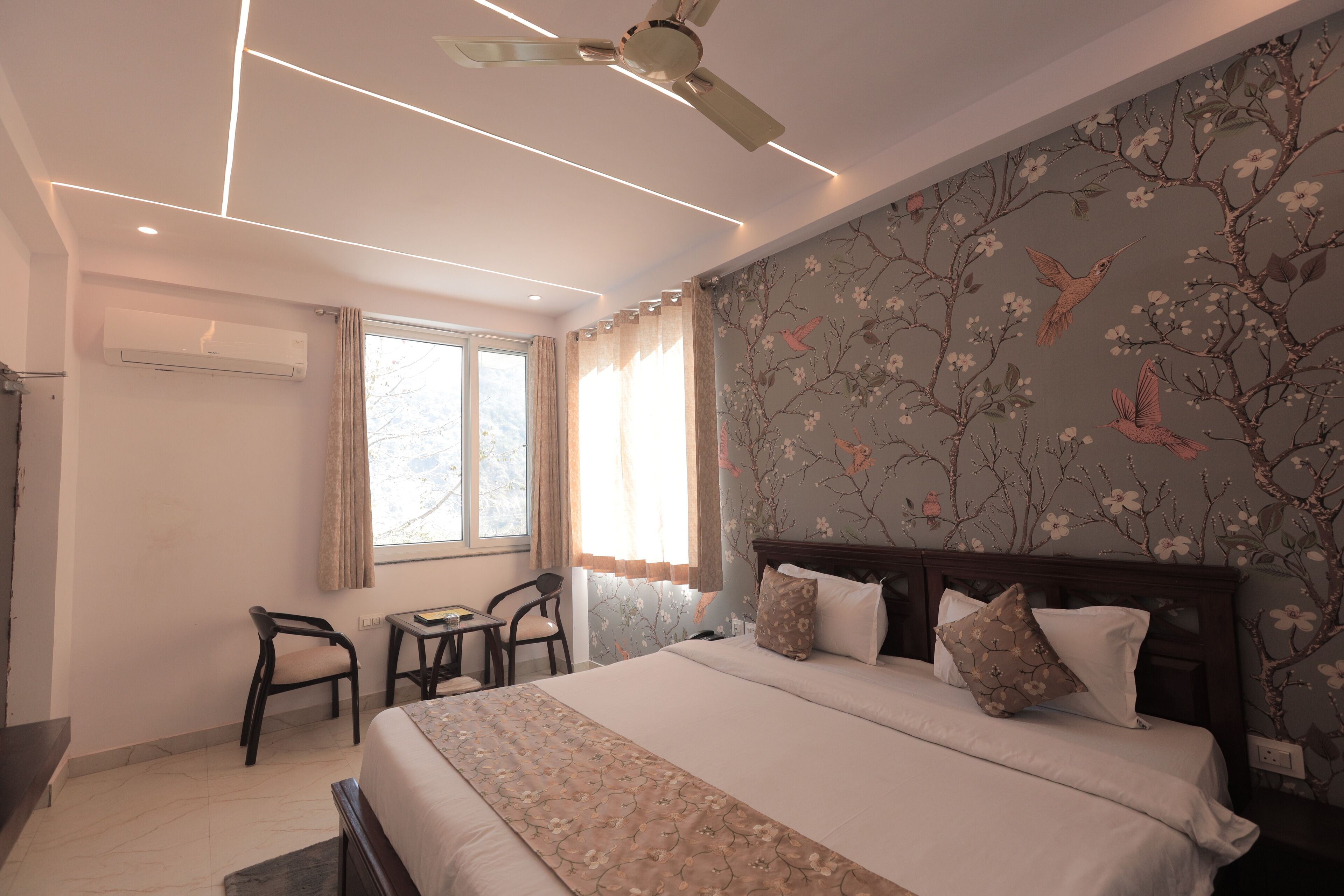 A bedroom with a bed, a desk, and a chair. There is a window with curtains, a ceiling fan, and a wall with floral wallpaper.