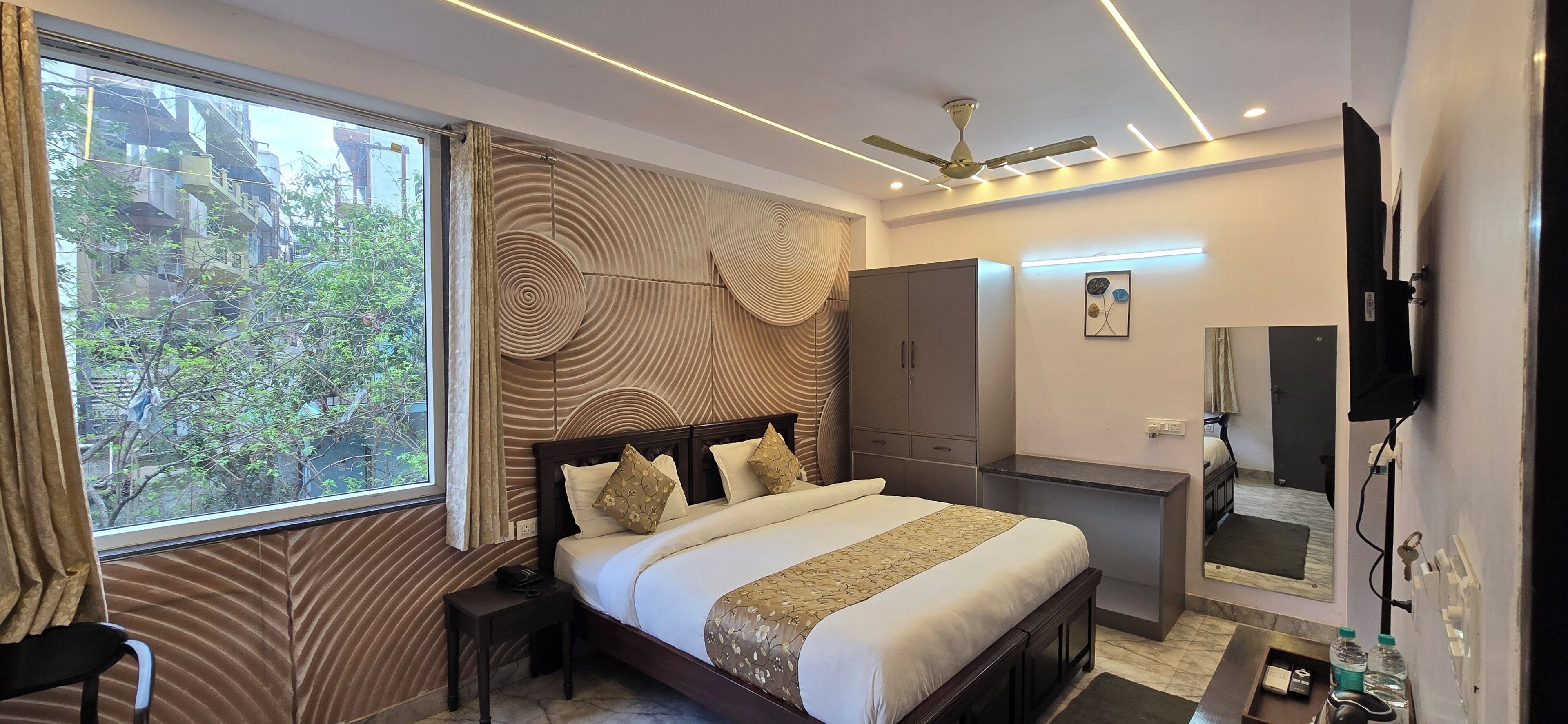 Ein modernes Hotelzimmer mit einem großen Bett, einem Kleiderschrank, einem Schminktisch mit Spiegel und einem Deckenventilator.