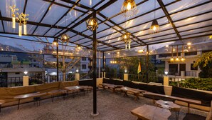 Een buitenruimte met tafels en banken, een pergola en hangende lampen.