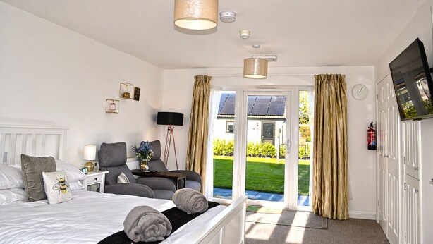 A bedroom with a bed, bedside table, lamp, and a view of the outdoors. - Lodge 5 - Lytton Tree Lodge, Reydon, Southwold (Reydon)