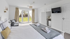A hotel room with a large bed, a TV mounted on the wall, a seating area with a table and chairs, and a view of the outdoors through a sliding door. - Lodge 5 - Lytton Tree Lodge, Reydon, Southwold (Reydon)