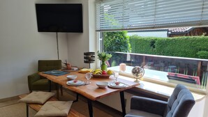 A dining area with a wooden table set for two, a green chair, a grey chair, and a bench with cushions. A flat-screen TV is mounted on the wall, and a window with blinds is visible. - Central in August 2022 newly renovated apartment Katja II with Kramerblick (Garmisch-Partenkirchen)