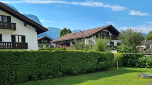 A well-maintained garden with a neatly trimmed hedge, a wooden deck, and a clear view of mountain ranges in the background. - Central in August 2022 newly renovated apartment Katja II with Kramerblick (Garmisch-Partenkirchen)