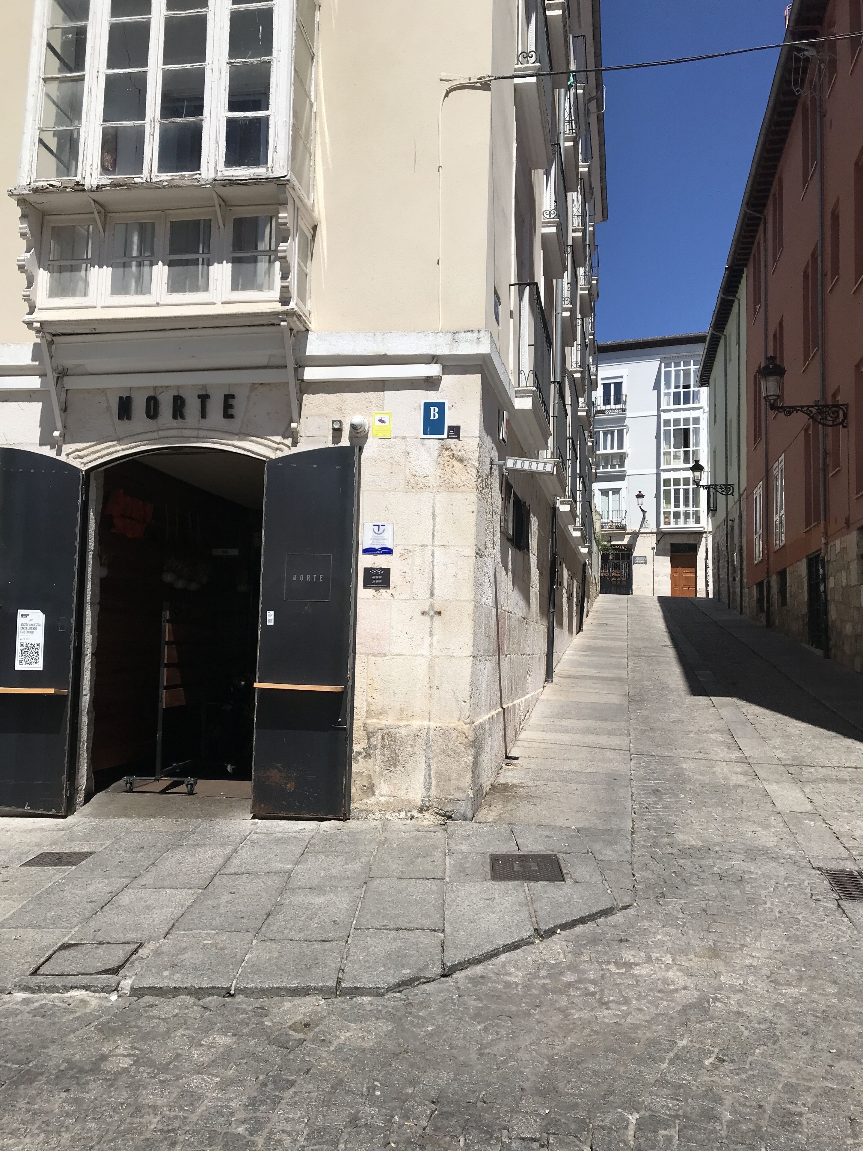 Una calle estrecha con edificios a ambos lados, un cartel que dice "MORTE" sobre una puerta, y una señal azul con una 'B' blanca en la pared.