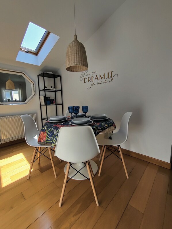 Un coin repas avec une table dressée pour quatre personnes, un luminaire suspendu en osier, une étagère murale avec des livres et une citation inspirante.