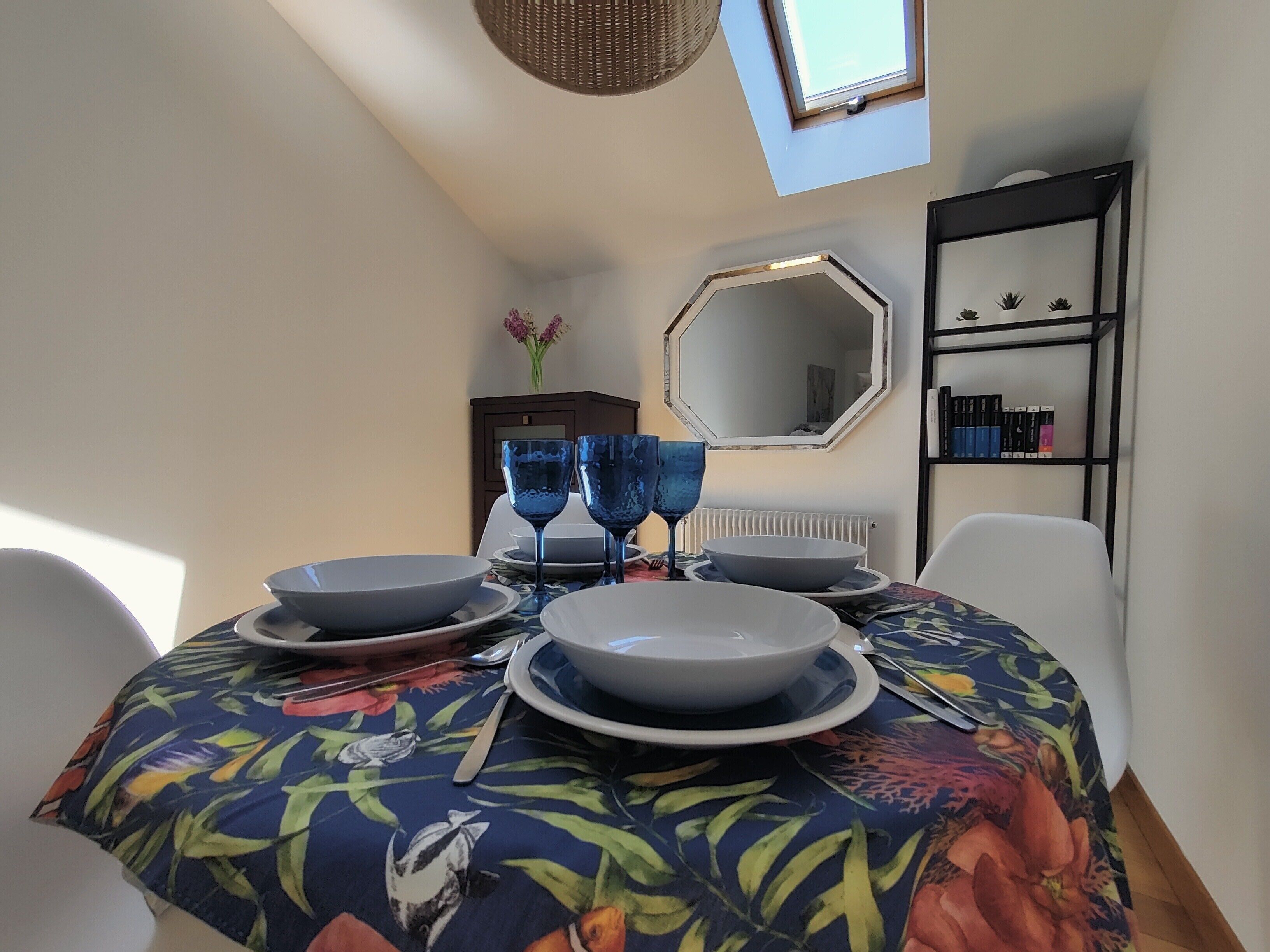 Uma mesa de jantar com copos e pratos azuis, uma toalha de mesa florida e uma estante com livros e objetos decorativos.