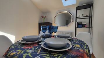 Uma mesa de jantar com copos e pratos azuis, uma toalha de mesa florida e uma estante com livros e objetos decorativos.