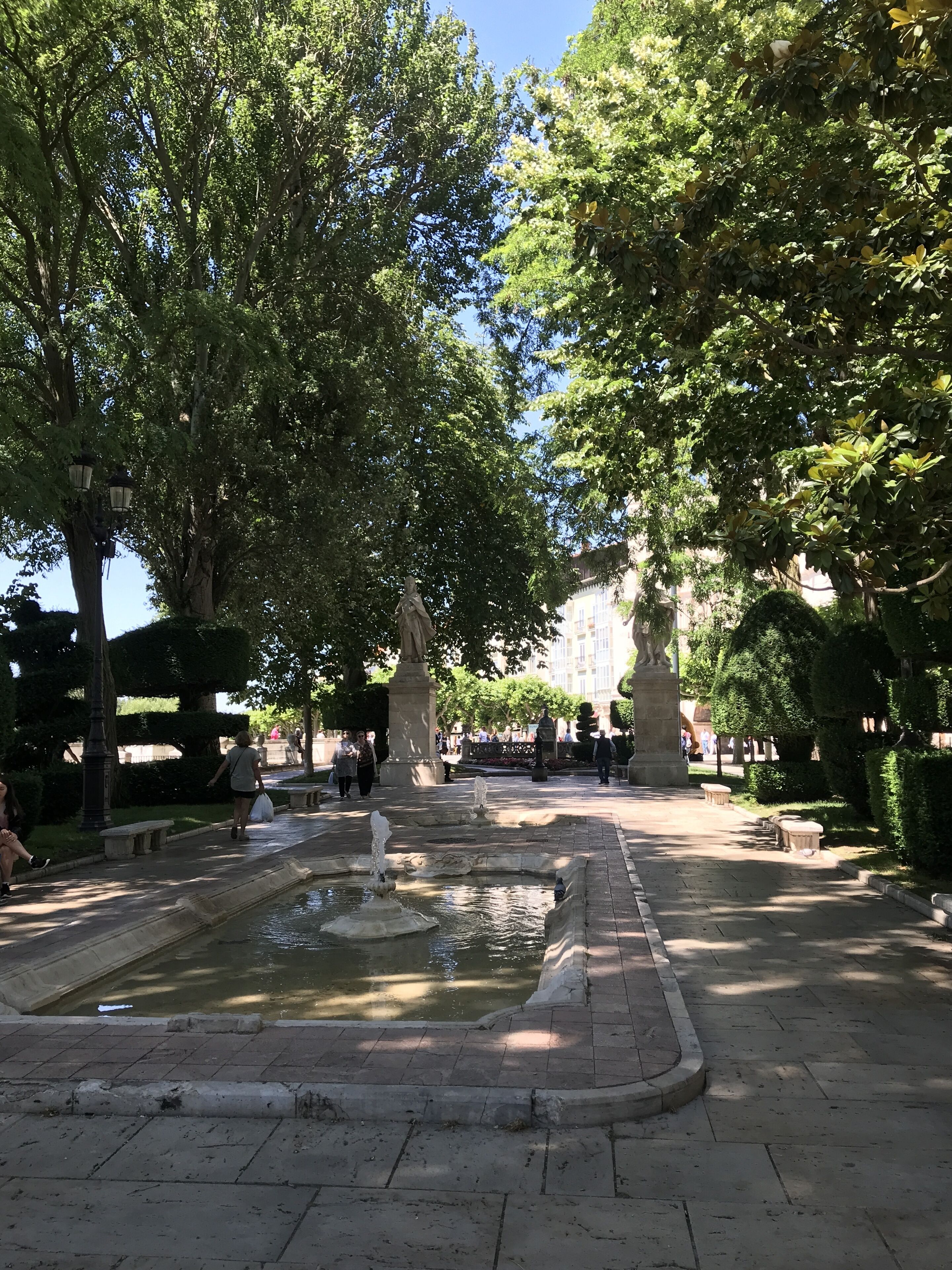 Une place publique agrémentée d’une fontaine, de bancs et de statues.