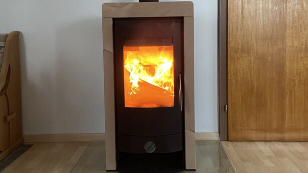 A wood-burning stove with a glass front, a black exhaust pipe, and a wooden door in the background. - Spacious, first floor vacation apartment in the Allgäu  (Memmingen)