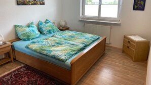 A bedroom with a wooden bed, a nightstand, a lamp, a radiator, a window, and a small wooden cabinet. - Spacious, first floor vacation apartment in the Allgäu  (Memmingen)