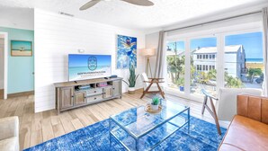 A modern living room with a large glass coffee table, a ceiling fan, a TV on a wooden cabinet, and a view of the ocean through large windows. - Gertrude's Siesta | Oceanview, 12 Steps to Beach, 4min Trolley to Village, Free Gear (Siesta Key)