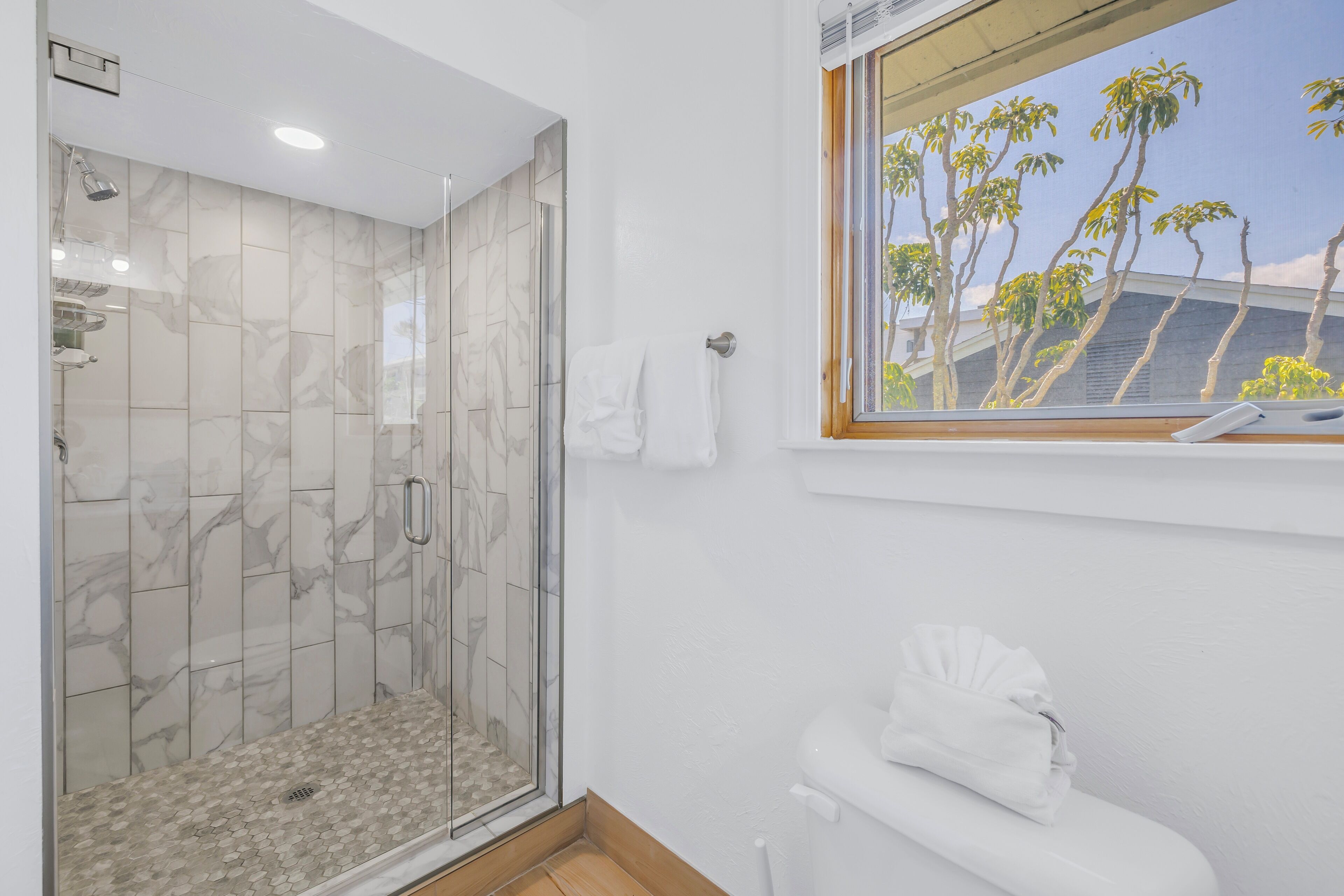 Un baño moderno con ducha envuelta en vidrio, inodoro y ventana con vista a palmeras.