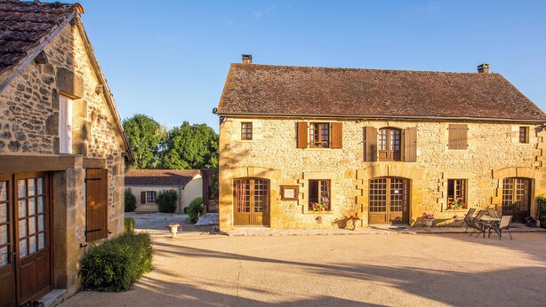 Un bâtiment en pierre doté de nombreuses fenêtres et portes, une cour avec une table et des chaises, et un petit jardin.