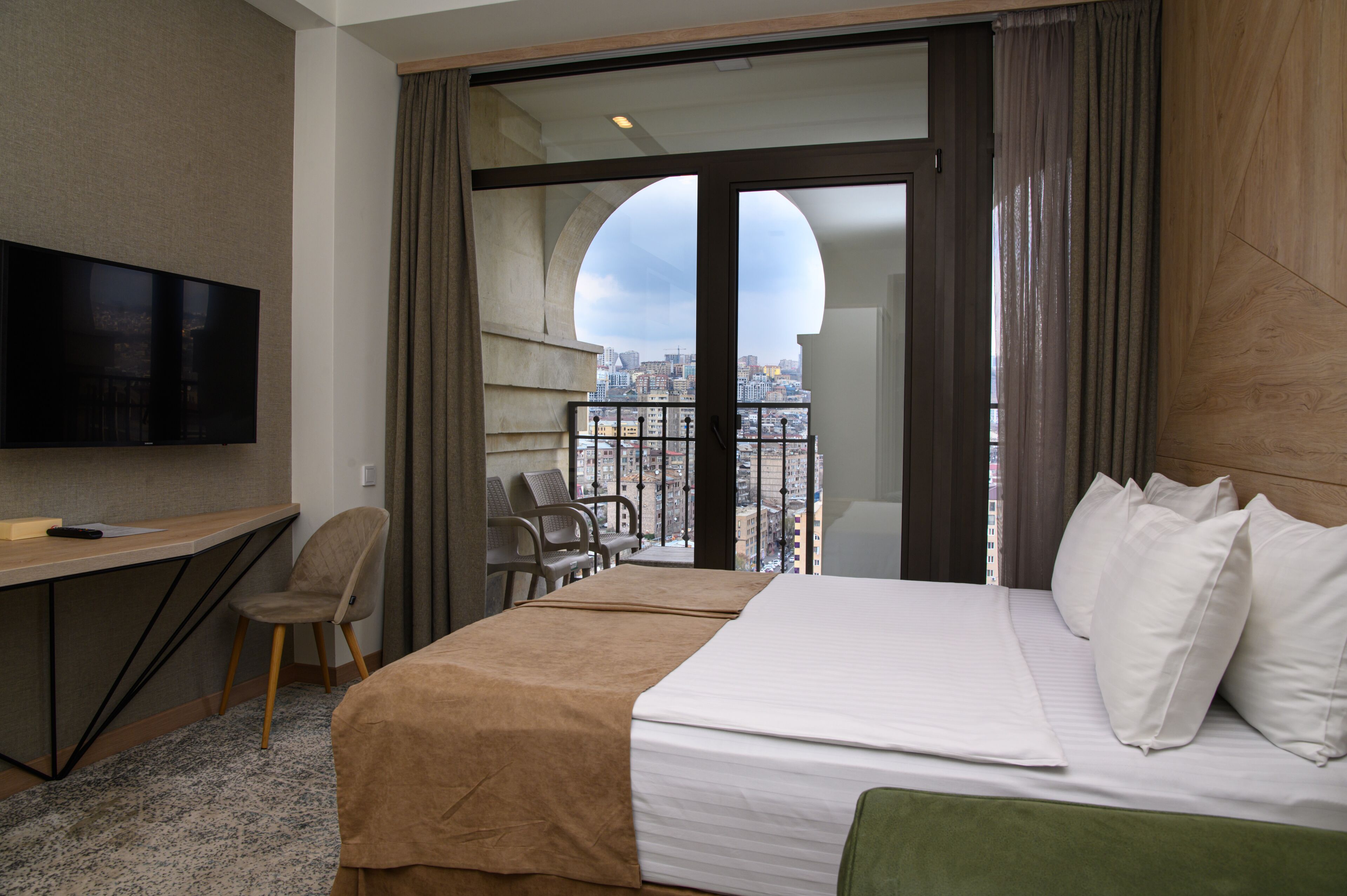 A hotel room with a bed, a desk with a television, a chair, and a balcony with a city view.