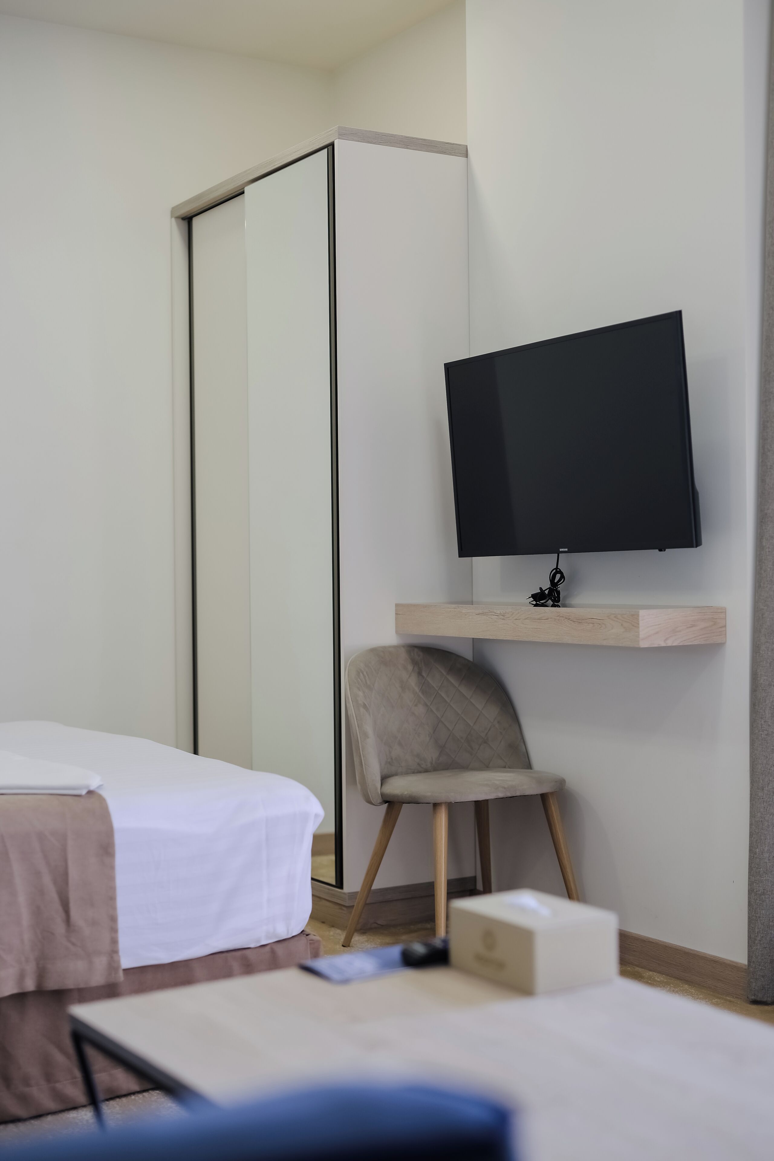 A hotel room with a bed, a desk with a television, a chair, and a small table.