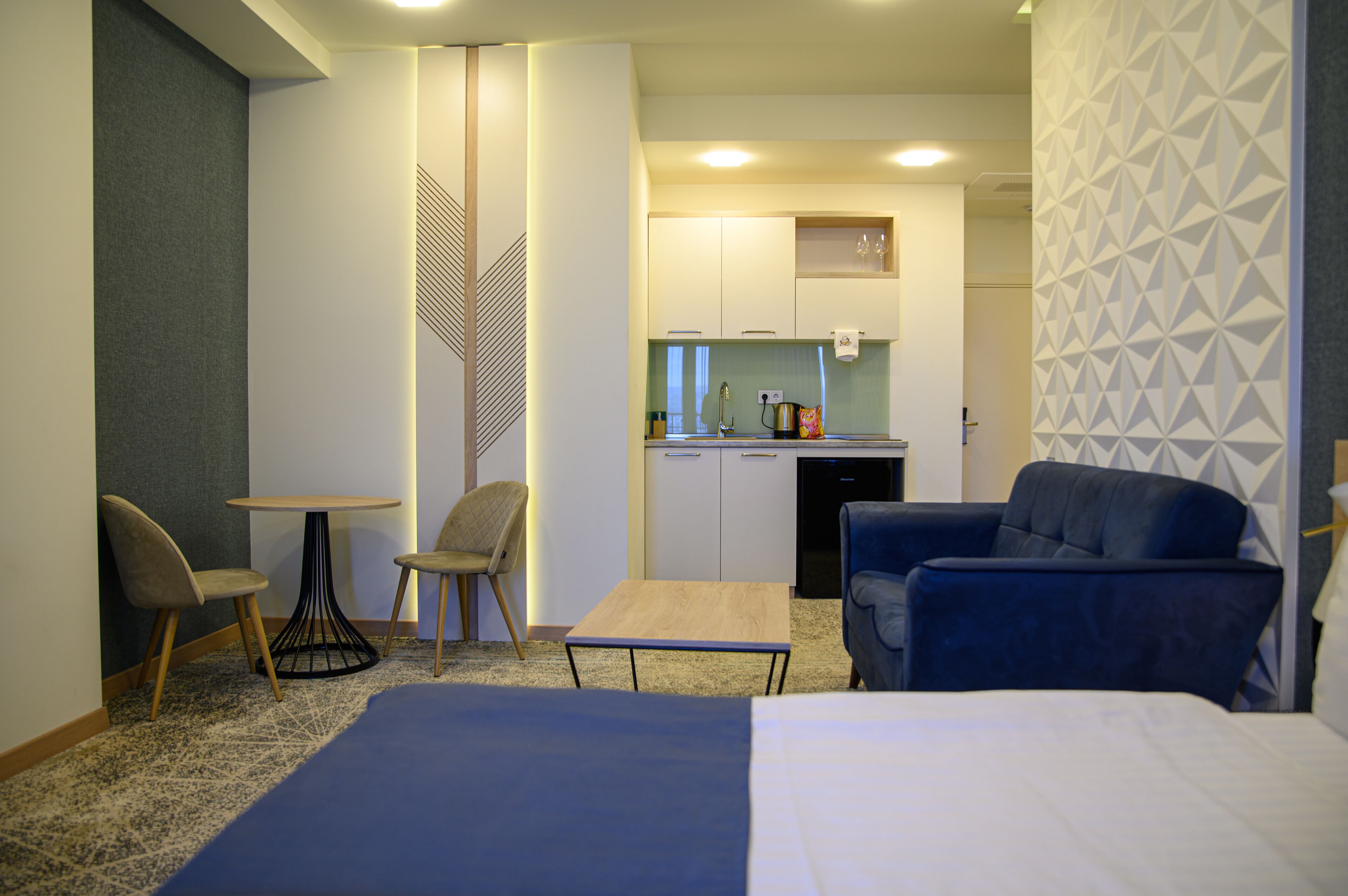 Habitación de hotel con cama, escritorio, dos sillas, una mesita y una kitchenette al fondo.