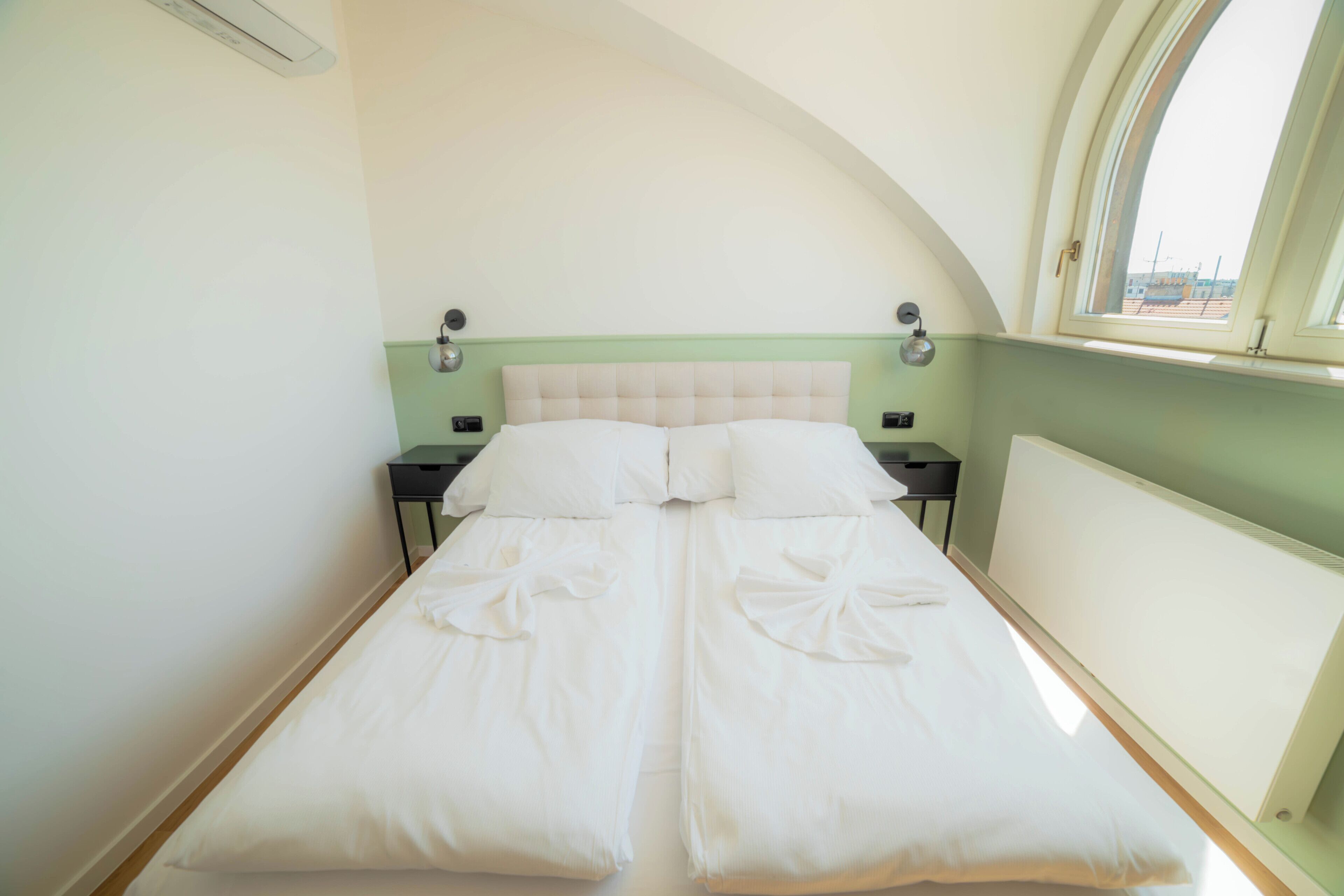 Cama doble con ropa blanca y cabecera verde en una habitación con tragaluz y vistas al exterior.