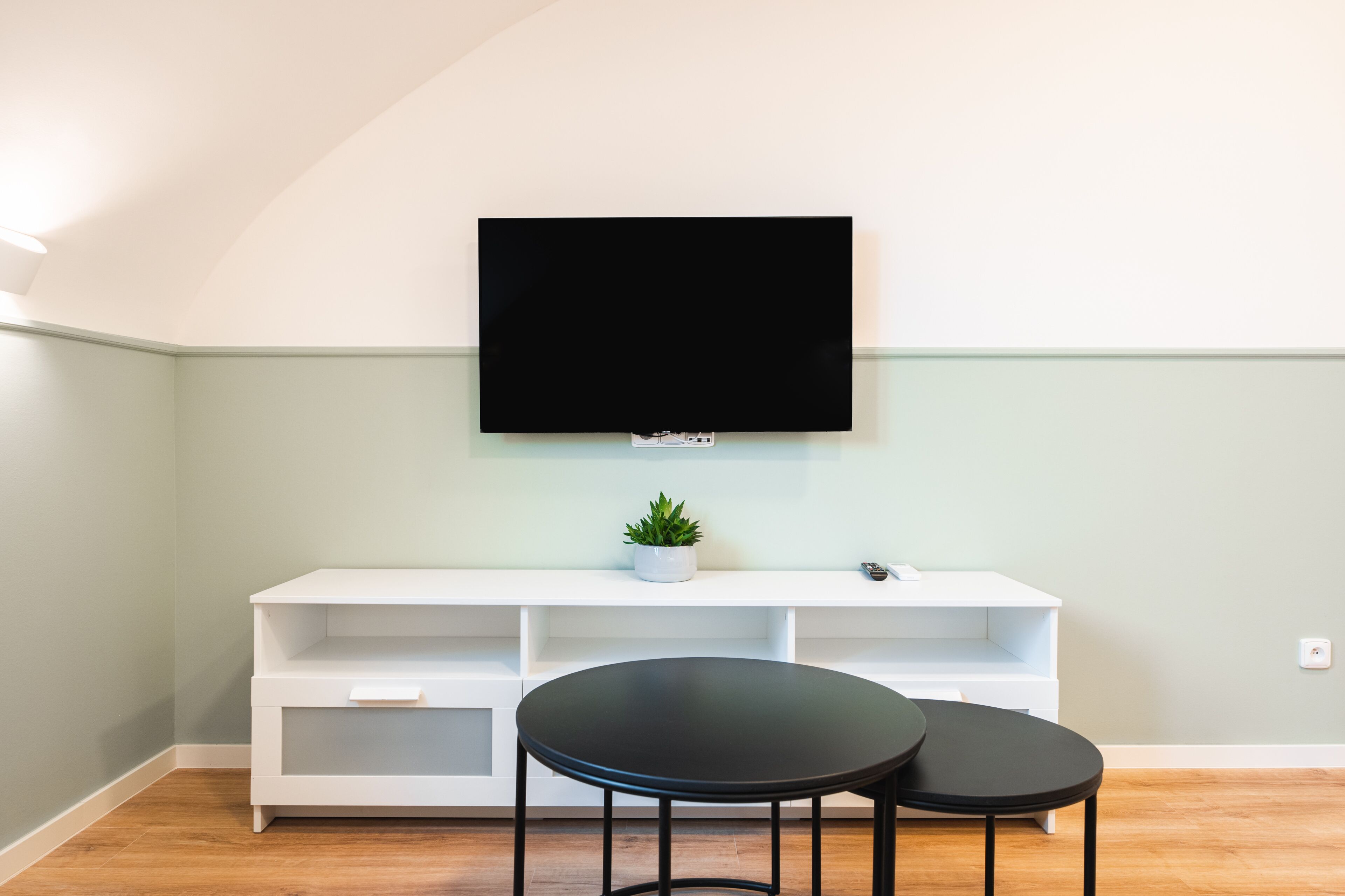 Una sala de estar moderna con un televisor de pantalla plana montado en la pared, un mueble blanco de entretenimiento debajo y dos mesas redondas negras.