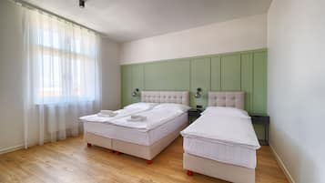A bedroom with two beds, a green headboard, a bedside table with a lamp, and a window with sheer curtains.
