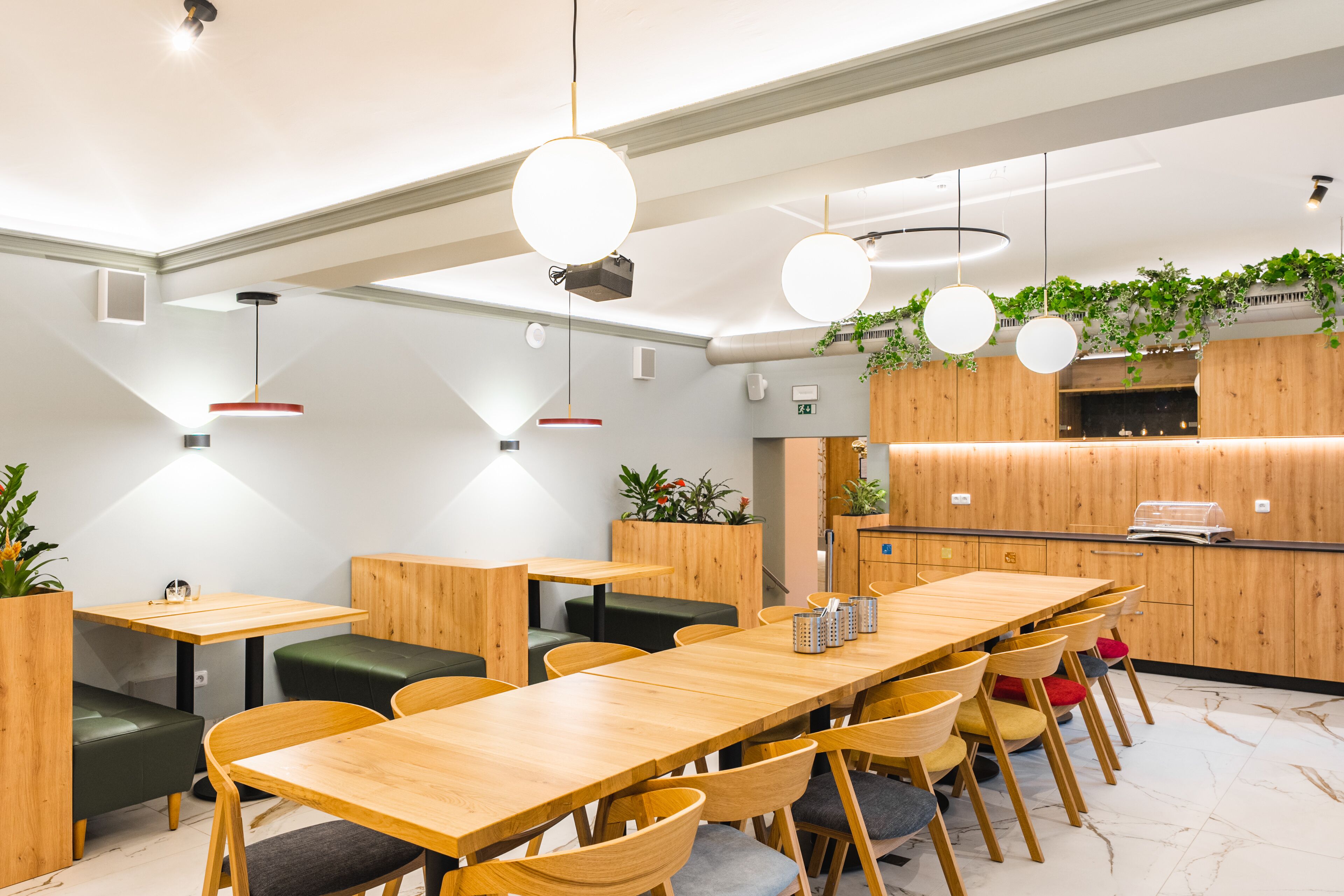 A modern dining area with wooden tables and chairs, pendant lighting, and a kitchen area with wooden cabinets and plants.
