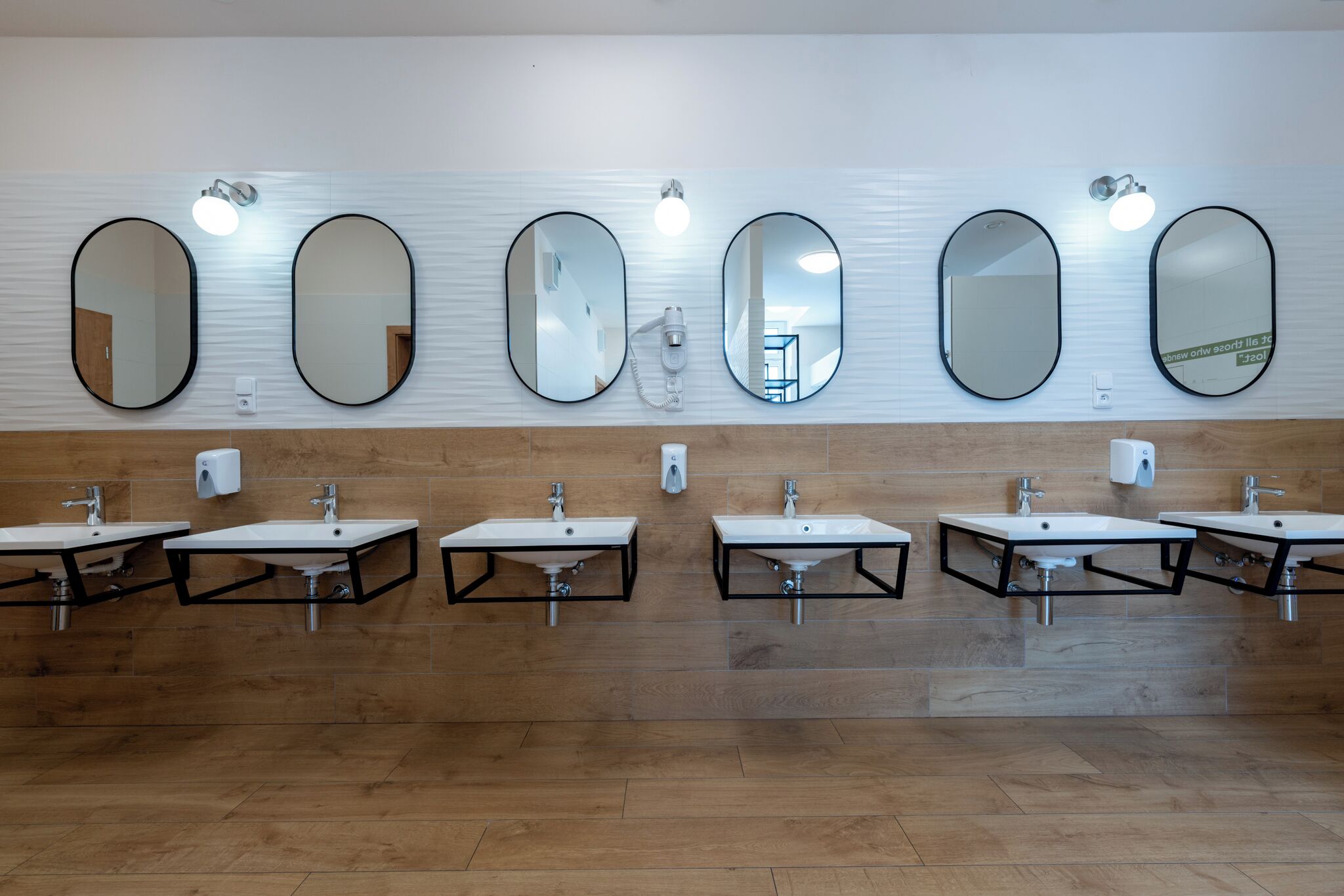 Een openbaar toilet met zes wasbakken, elk met een spiegel erboven. Er hangen wandlampen boven de spiegels.