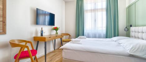 Quarto de hotel com uma cama, uma escrivaninha, uma cadeira, uma televisão e uma janela com cortinas.