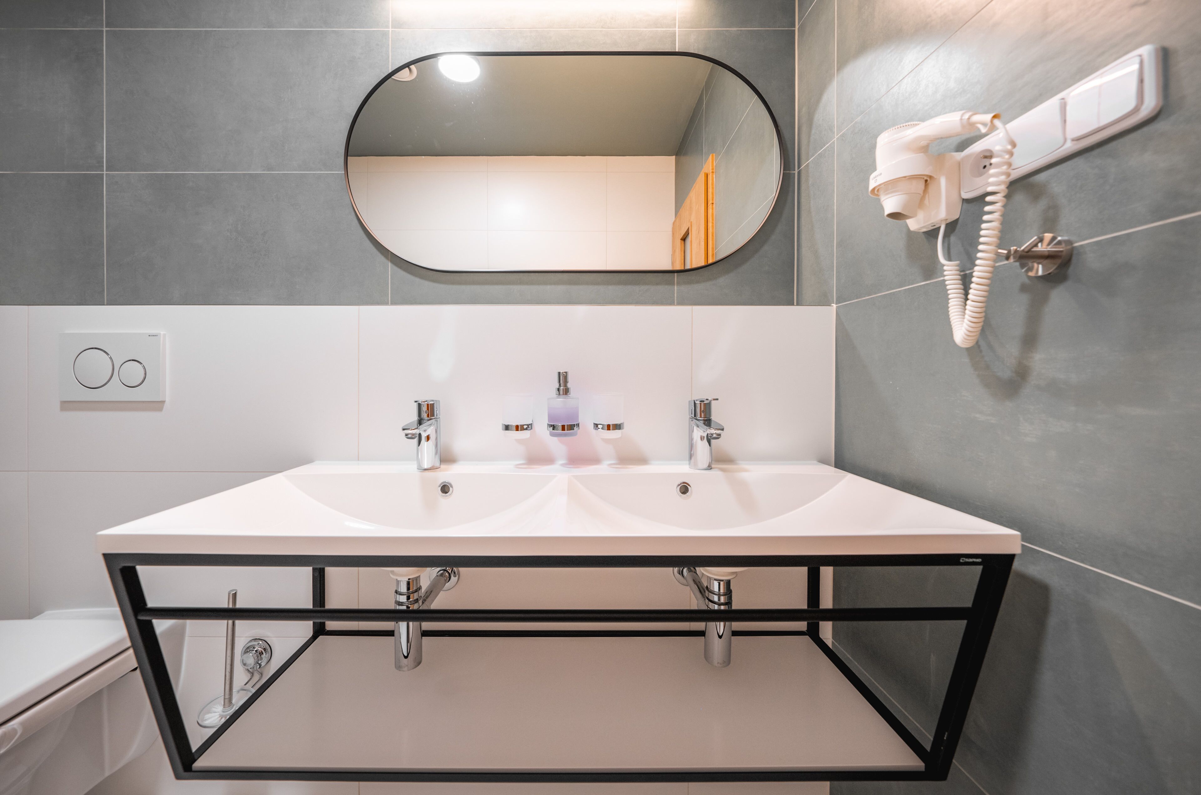 Un baño moderno con un lavabo blanco, dos grifos, un espejo y un secador de pelo montado en la pared.