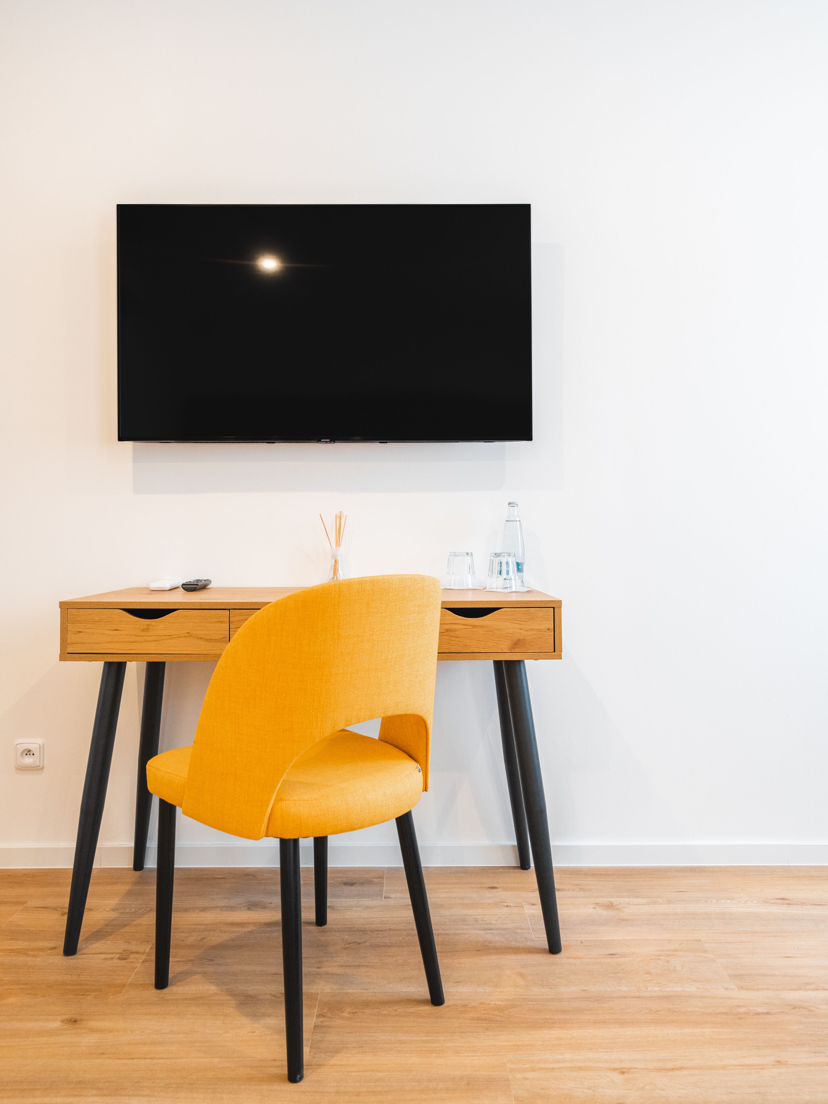 Une chambre moderne avec un bureau en bois, une chaise jaune, un téléviseur à écran plat fixé au mur et une petite table sur laquelle se trouvent un verre et une bouteille.