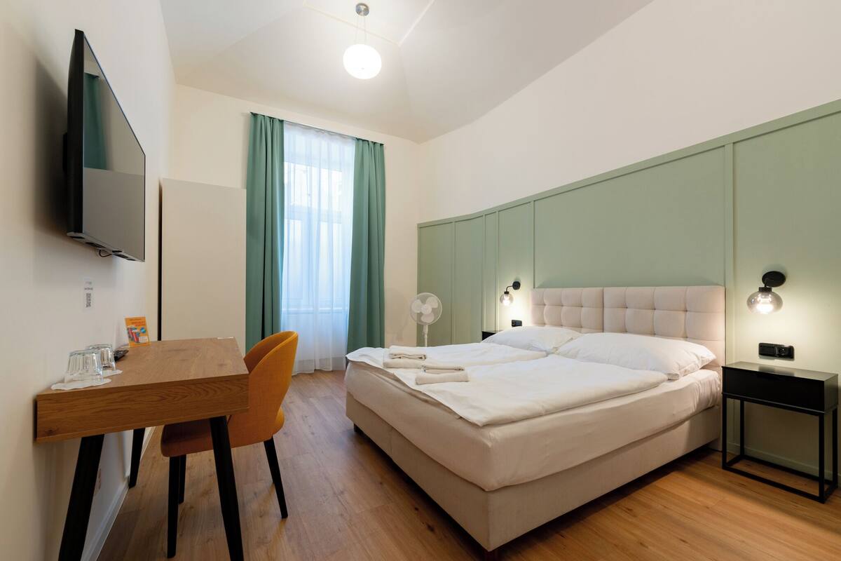 A hotel room with a bed, a desk with a chair, a television, and a window with curtains.