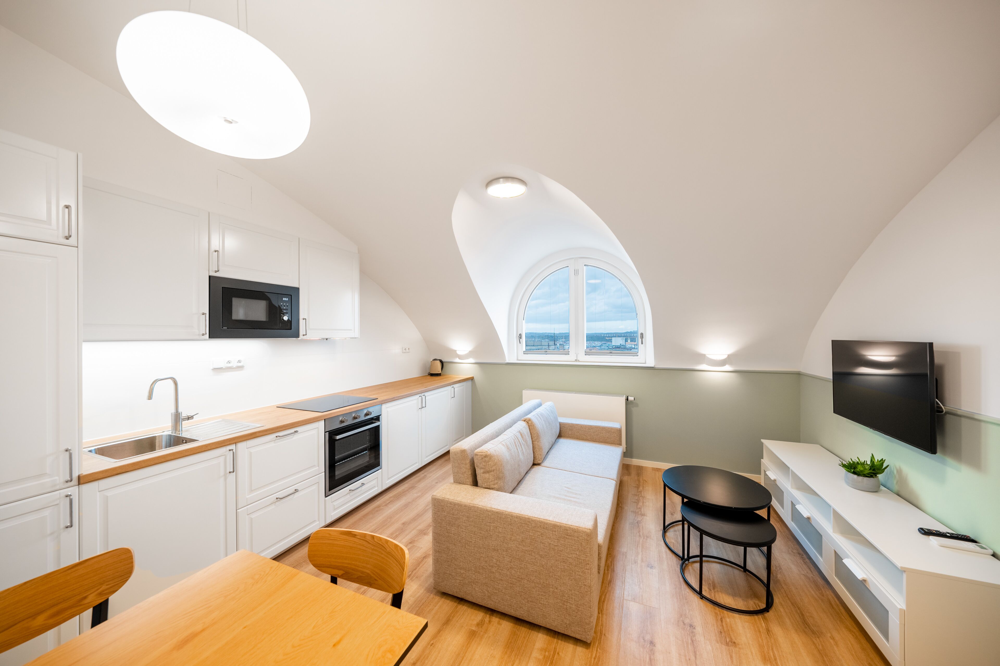 Una cocina moderna con armarios blancos, un microondas y una encimera de madera. Hay un sofá y una mesa de comedor con sillas. Un televisor está montado en la pared, y una ventana ofrece vistas a la ciudad.